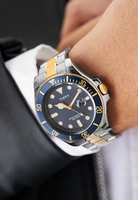 Montre Timex avec un cadran bleu, index dorés lumineux, bracelet en acier inoxydable et ton or, étanche jusqu'à 100m, fenêtre de date à 3 heures.
