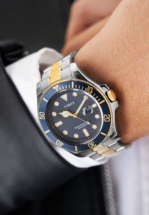 Montre Timex avec un cadran bleu, index dorés lumineux, bracelet en acier inoxydable et ton or, étanche jusqu'à 100m, fenêtre de date à 3 heures.