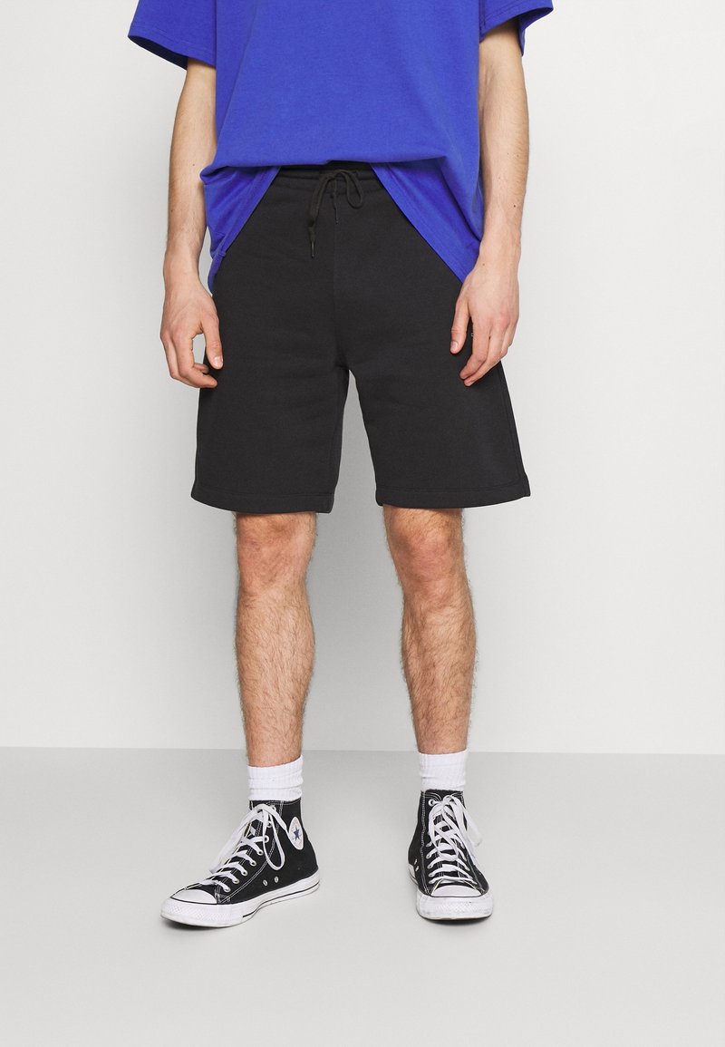Homme portant un short noir à cordon, des baskets montantes noires avec des lacets blancs, des chaussettes blanches et une chemise bleue, debout devant un fond uni.