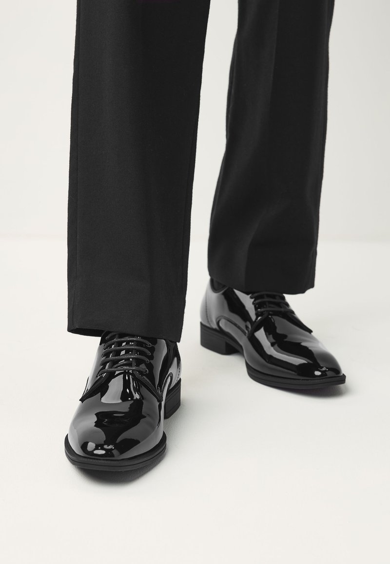 Chaussures de ville brillantes noires portées avec un pantalon noir sur un fond clair.