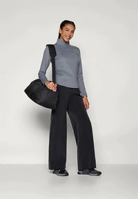 Gray long-sleeve top, black wide-leg pants, and black sneakers. Model holds a black duffel bag. Smooth texture, athletic design.