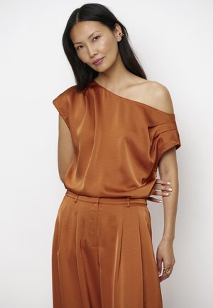 Vrouw met lang donker haar, gekleed in een roestkleurige off-shoulder satijnen top en bijpassende high-waisted wijde broek, poserend tegen een witte achtergrond.