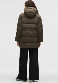 Kurtka puchowa w kolorze oliwkowej zieleni z kapturem, pikowanym wzorem i szerokimi rękawami. Noszona na czarnych, luźnych spodniach oraz zielonkawym obuwiu.