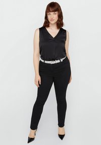 Top senza maniche in satin nero con scollatura a V, abbinato a jeans skinny neri e una cintura argentata scintillante, accentuato da tacchi neri a punta.