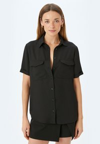 Camisa negra de manga corta hecha de tela suave. Presenta un cierre de botones en la parte delantera, cuello y dos bolsillos en el pecho. Diseño simple y ajustado.