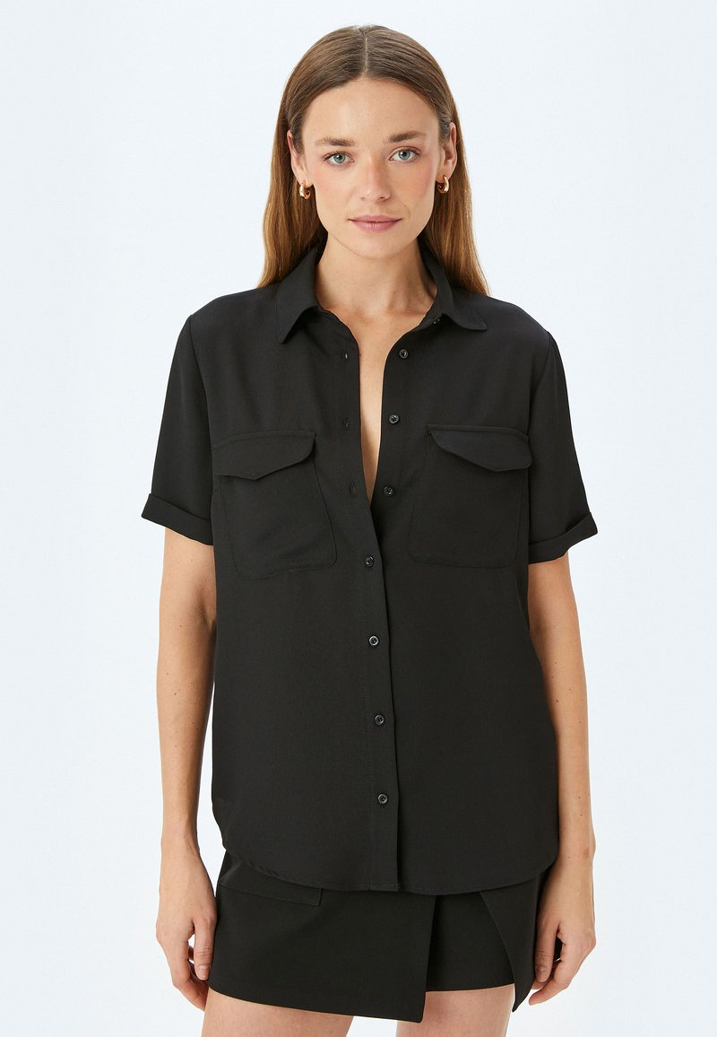 Camisa negra de manga corta hecha de tela suave. Presenta un cierre de botones en la parte delantera, cuello y dos bolsillos en el pecho. Diseño simple y ajustado.
