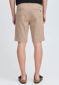 Pantalones cortos de algodón beige con corte recto, dos bolsillos traseros y dobladillo limpio. Llevados con sandalias marrones de tipo slide.