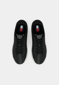 Een paar zwarte lage sneakers met witte zolen, zwarte veters en het Tommy Jeans-logo op de tongen en binnenzolen, gezien van boven.