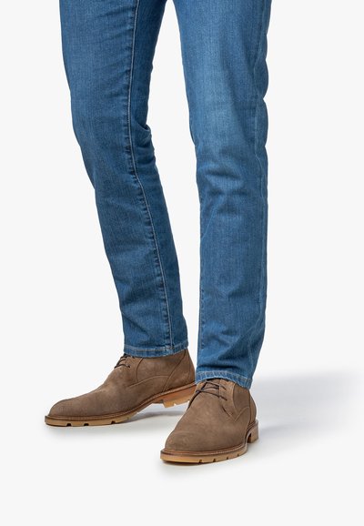 Man wearing brown suede lace-up boots and blue slim-fit jeans standing on a white background.