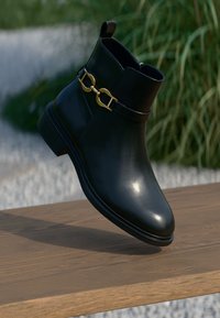 Schwarze Leder-Stiefelette mit glatter Oberfläche, dekoriert mit einer goldenen Kette und einem niedrigen Blockabsatz. Auf einer Holzoberfläche platziert.