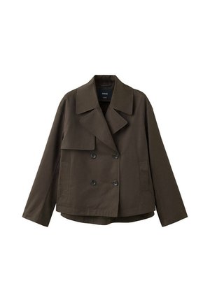 Mujer joven con chaqueta cruzada de color marrón oscuro, suéter verde claro, vaqueros de pierna ancha azul claro y zapatos oscuros, de pie sobre un fondo blanco.
