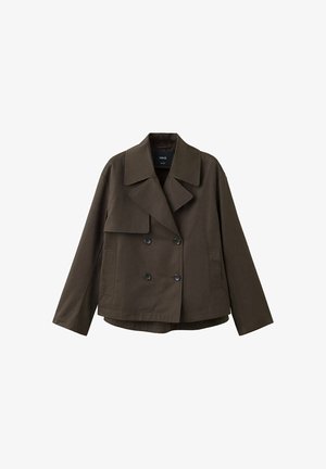 Jeune femme portant une veste croisée marron foncé, un pull vert clair, un jean bleu clair à jambes larges et des chaussures foncées, debout sur un fond blanc.
