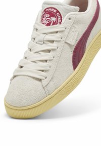Bijele suede tenisice s bordo naglascima, ravnim vezicama i strukturiranim gumennim potplatom. Imaju logotip na jeziku i izraženu bočnu prugu.