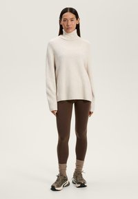 Maglione a collo alto beige chiaro, vestibilità ampia, polsini a coste; abbinato a leggings marroni e calzini alla caviglia texture, completato da sneakers sportive.