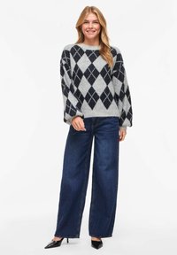 Pull en maille à losanges gris et marine avec manches larges, associé à un jean pattes d'éléphant bleu marine taille haute et des escarpins noirs à bouts pointus.