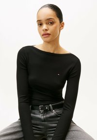 Maglia nera a coste con maniche lunghe e scollatura ampia, con piccolo logo sul petto, abbinata a jeans grigi in denim e una cintura nera.