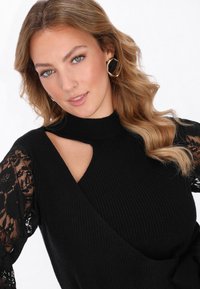 Czarny prążkowany top z dzianiny z półgolfem i otwartym designem na ramionach. Długie koronkowe rękawy dodają tekstury dzięki kwiatowym wzorom.