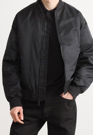 Personne portant une veste bomber noire zippée sur une chemise noire à col et un pantalon noir, une main dans la poche et l'autre détendue.