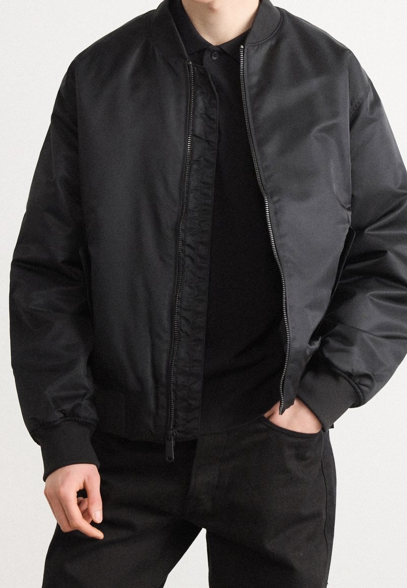 Personne portant une veste bomber noire zippée sur une chemise noire à col et un pantalon noir, une main dans la poche et l'autre détendue.