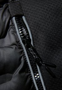 Svart tyg med en slät textur, med en dragkedja med glansig finish och grå detaljer. Dragkedjan är delvis öppen.