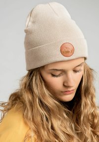 Jasnobeżowa wełniana czapka z podwinięciem, z okrągłą skórzaną naszywką przedstawiającą góry i drzewa na teksturowanym tle.