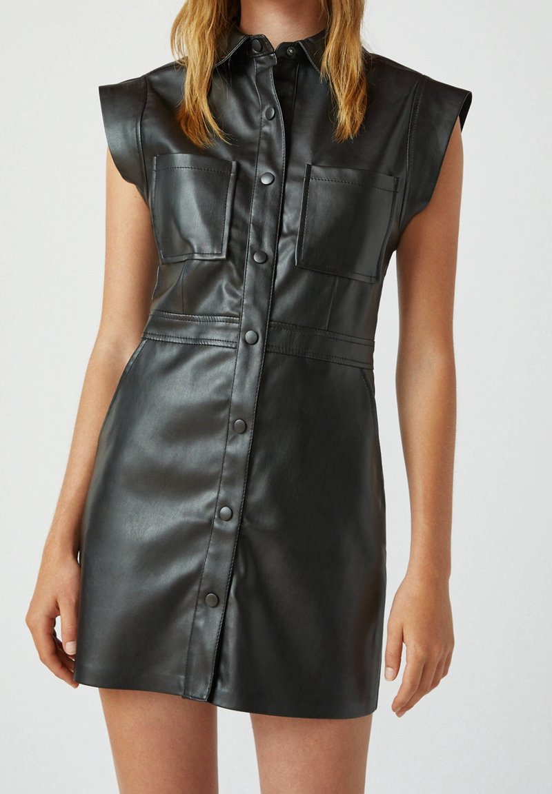 Robe noire sans manches en simili cuir avec boutons-pression à l'avant, deux poches poitrine et épaules structurées portée par un mannequin sur fond uni.