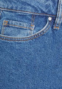 Detailní záběr na denní látku v světlomodré barvě, s výrazným oranžovým prošitím a malou přední kapsičkou s kovovým nýtem.