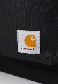 Bolsa de tela negra con una etiqueta blanca que presenta un logo en naranja y blanco, además del nombre de la marca "carhartt" impreso en color burdeos.
