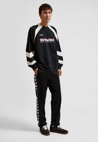 Young man standing wearing black and white "Swarm" sweatshirt, black track pants with white chevron stripes, and black sneakers.