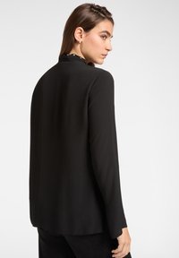 Blusa nera a maniche lunghe realizzata in tessuto leggero con una vestibilità dritta. Presenta un colletto rotondo e dettagli accentuati sottili alla scollatura.