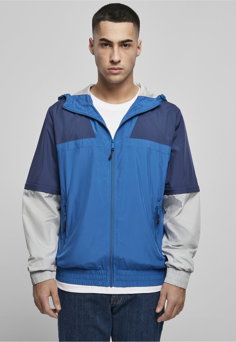 Joven con chaqueta con capucha con cremallera en azul, azul marino y gris claro sobre una camiseta blanca, de pie frente a un fondo gris liso.