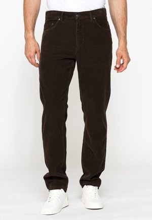 Uomo che indossa pantaloni in velluto a coste marrone scuro e sneakers bianche, in piedi su sfondo bianco.