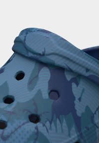 Gros plan sur une sandale sabot à motif camouflage bleu et bleu-vert avec des trous de ventilation et une bride au talon.