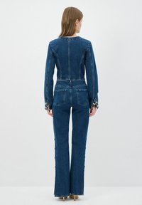Mono de mezclilla en azul oscuro, con un cuerpo ajustado, piernas acampanadas, puños abotonados y bolsillos traseros. Textura suave y diseño estructurado.