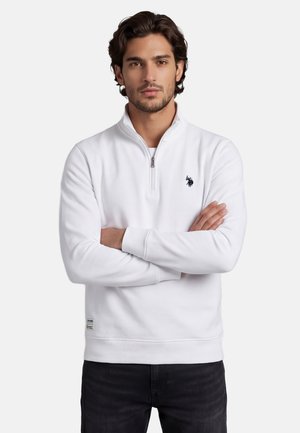 Mann mit verschränkten Armen, der ein weißes Viertel-Zip-Sweatshirt mit kleinem schwarzem besticktem Logo auf der Brust und schwarze Hose trägt.