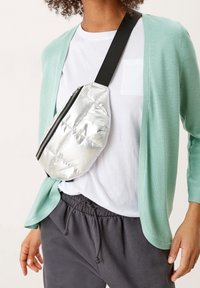 Silver quiltad midjeväska med en övre dragkedja och svart rem, bärs över en vit T-shirt under en mintgrön cardigan och grå sweatpants.