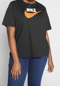 Černé bavlněné tričko s krátkým rukávem s bílým logem "NIKE" a oranžovým designem swoosh. Volný střih s kulatým výstřihem.