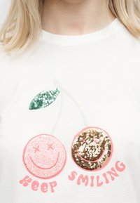 T-shirt avec deux cerises en sequins, l'une rose et l'autre dorée, toutes deux avec des visages souriants, et un texte rose en dessous disant "Keep Smiling".
