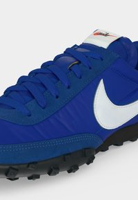 Μπλε αθλητικό παπούτσι με λευκό λογότυπο Nike, υφασμένη μαύρη σόλα και μπλε κορδόνια, εμφανίζεται από κοντινή πλευρική γωνία.