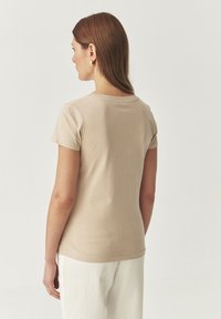 T-shirt beige ajusté à manches courtes, col rond, texture lisse et ourlet légèrement arrondi, porté avec un pantalon blanc.