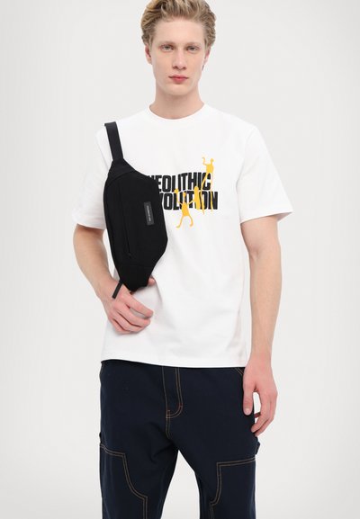 Geantă de umăr neagră cu o suprafață texturată, purtată peste un tricou alb cu grafice îndrăznețe în negru și galben. Îmbrăcăminte casual cu pantaloni bleumarin.