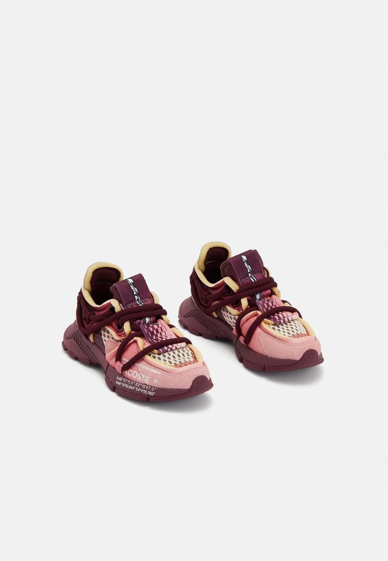 Par af burgundy og pink atletiksneakers med mesh- og ruskindsdetaljer, tykke såler og unikt snøresystem på en hvid baggrund.
