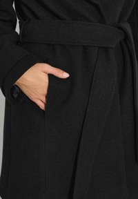 Ręka włożona w boczną kieszeń czarnego płaszcza z paskiem, wykonanego z fakturowanej tkaniny.