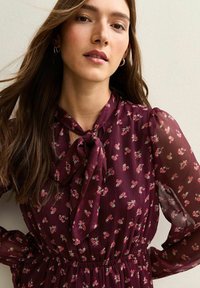 Vestido floral burdeos con mangas transparentes, detalle de cuello con lazo y cintura elástica, con pequeños patrones de flores rosas y blancas.