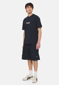 Námořnické tričko s logem "DICKIES", spárované s tmavými cargo šortkami s kapsami. Model má na sobě bílé tenisky a sportovní ponožky.