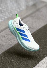 Weißer Sportschuh mit blauen und grünen Akzenten. Verfügt über ein glattes Obermaterial, eine abgerundete Schuhspitze und eine weiche, hellblaue Sohle. Rutschbewegung wird dargestellt.