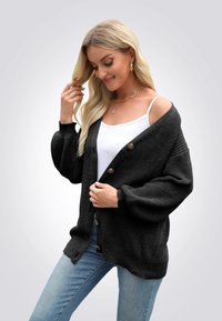 Černý pletený cardigan s velkými knoflíky, spadlými rameny a žebrovanými manžetami. Nosený přes bílý top bez rukávů, v kombinaci s modrými džínami.