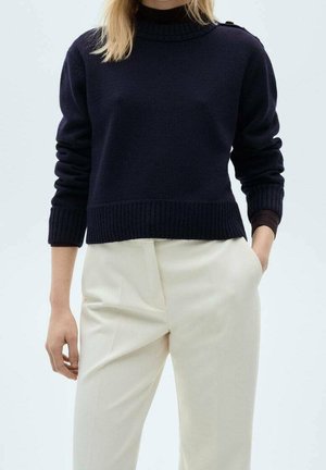 Personne portant un pull bleu marine foncé à manches longues et un pantalon crème taille haute, une main dans la poche et une main détendue sur le côté.