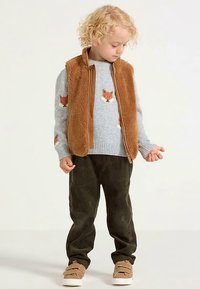 Enfant portant une veste en polaire marron par-dessus un pull gris avec des motifs de renard, associée à un pantalon en velours côtelé vert et des chaussures beige à enfiler.