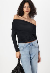 Melna off-the-shoulder blūze ar ručņu tekstūru, garām piedurknēm, apvienota ar augsto jostasvietu gaiši zilajiem džinsiem un melnu rakstainu somu.
